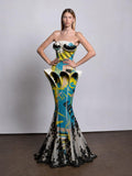 strapless mermaid gown with a sculptural corset-style bodice and flared skirt. The dress has a bold abstract print in turquoise, yellow, black, and off-white, with black lace at the hem.