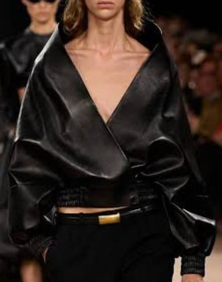 A runway photo of a model walking down a fashion show catwalk wearing a dramatic black satin-like wrap jacket, black trousers, black sunglasses, and carrying a black handbag.