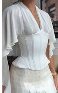 A woman wearing a white structured blouse with dramatic ruffled sleeves and a deep V-neckline, paired with a white textured mini skirt, shown in an indoor setting.