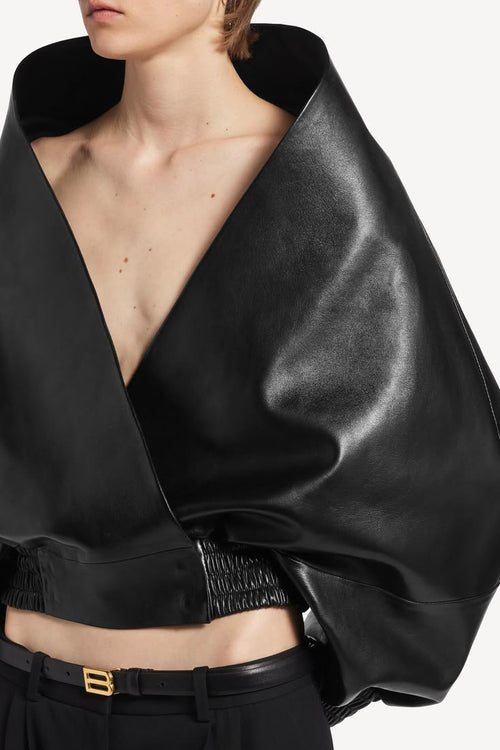 Close-up of a model wearing a black draped wrap top with a deep V-neck, wide voluminous sleeves, and a gathered waist, paired with black trousers and a black belt with a gold buckle.