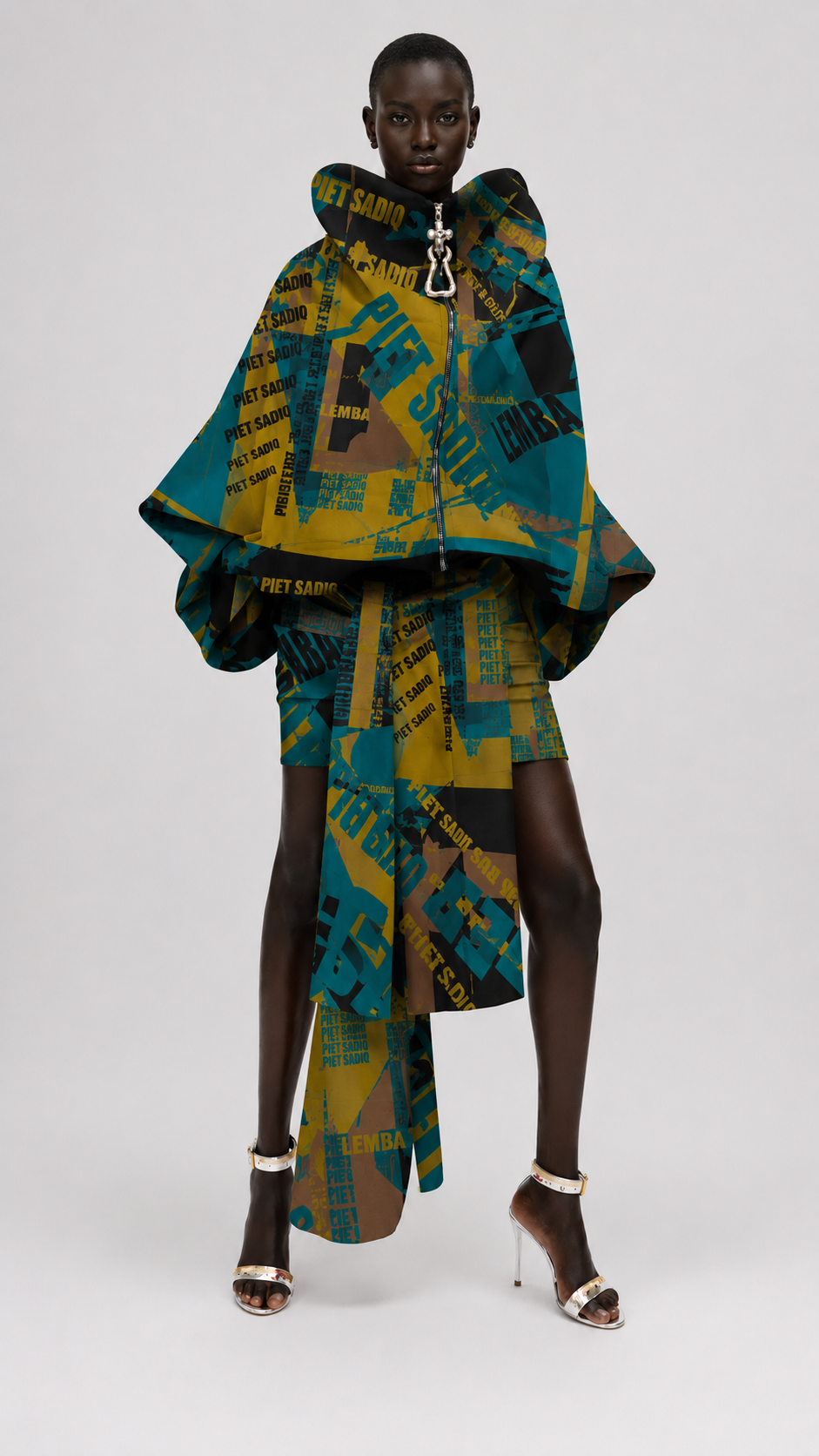 Oversized cape-style top and matching skirt in a bold abstract print. The garment features teal, mustard yellow, black, and tan sections with text-like graphics, and the model is wearing metallic heeled sandals.