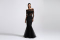 A model wearing a black off-the-shoulder evening gown with sheer long sleeves and a flared tulle hem, photographed against a light gray studio background.