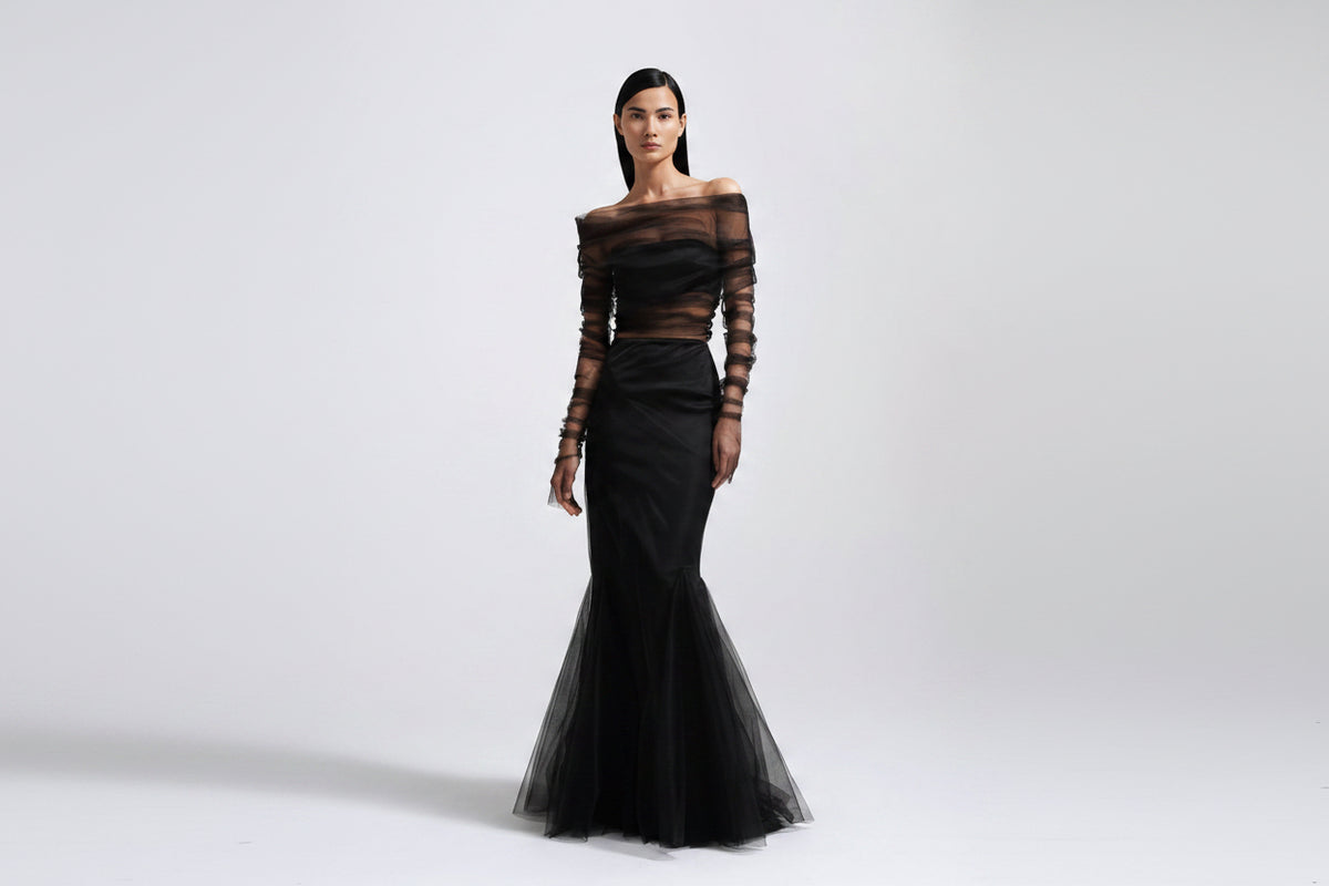 A model wearing a black off-the-shoulder evening gown with sheer long sleeves and a flared tulle hem, photographed against a light gray studio background.