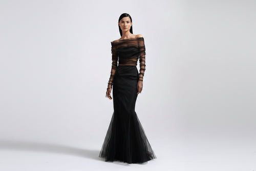 A model wearing a black off-the-shoulder evening gown with sheer long sleeves and a flared tulle hem, photographed against a light gray studio background.