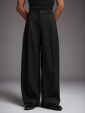 A model wearing high-waisted, wide-leg trousers in black with thin vertical pinstripes, paired with a black top and black leather shoes, set against a neutral gray studio background.