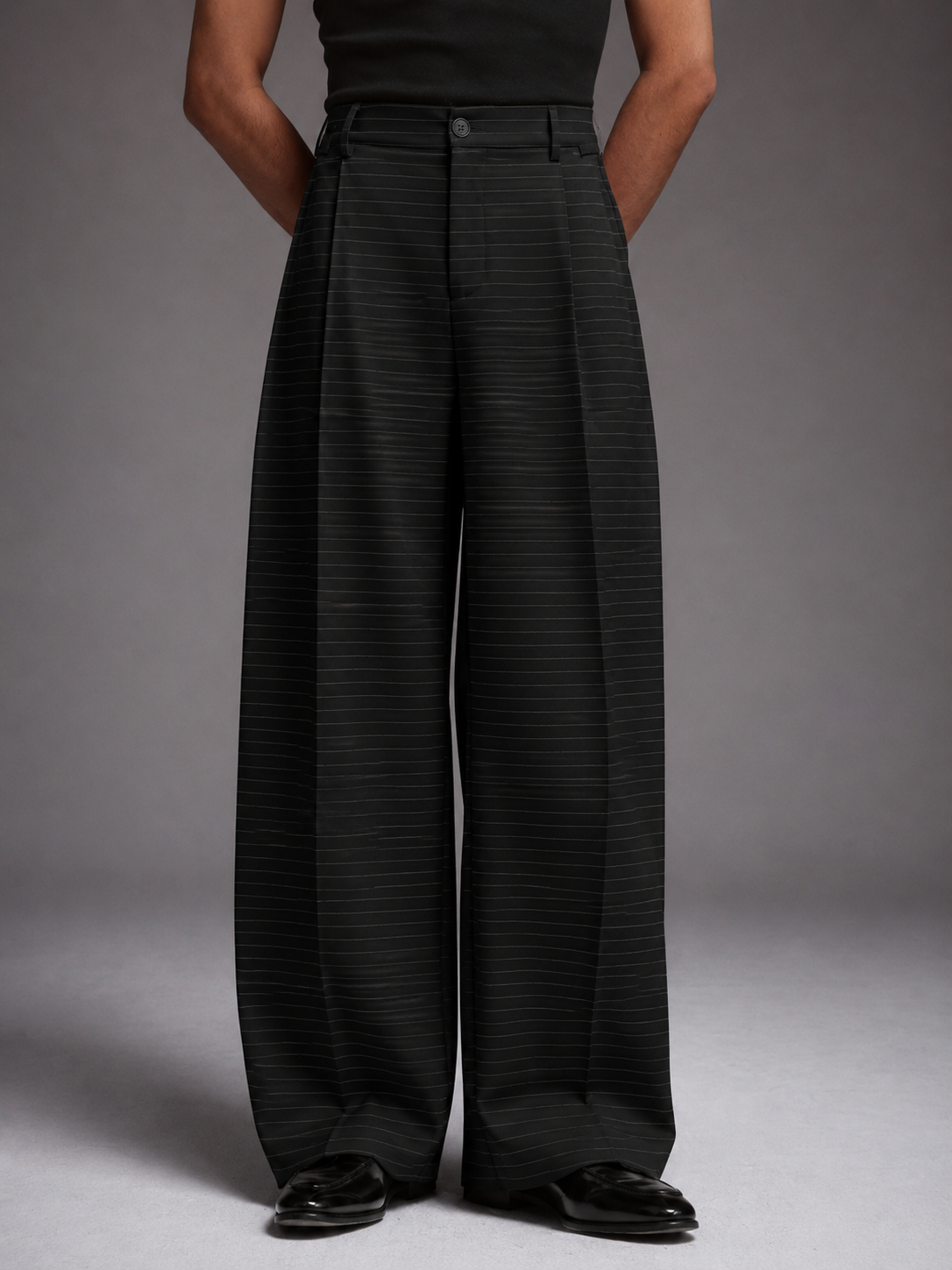 A model wearing high-waisted, wide-leg trousers in black with thin vertical pinstripes, paired with a black top and black leather shoes, set against a neutral gray studio background.